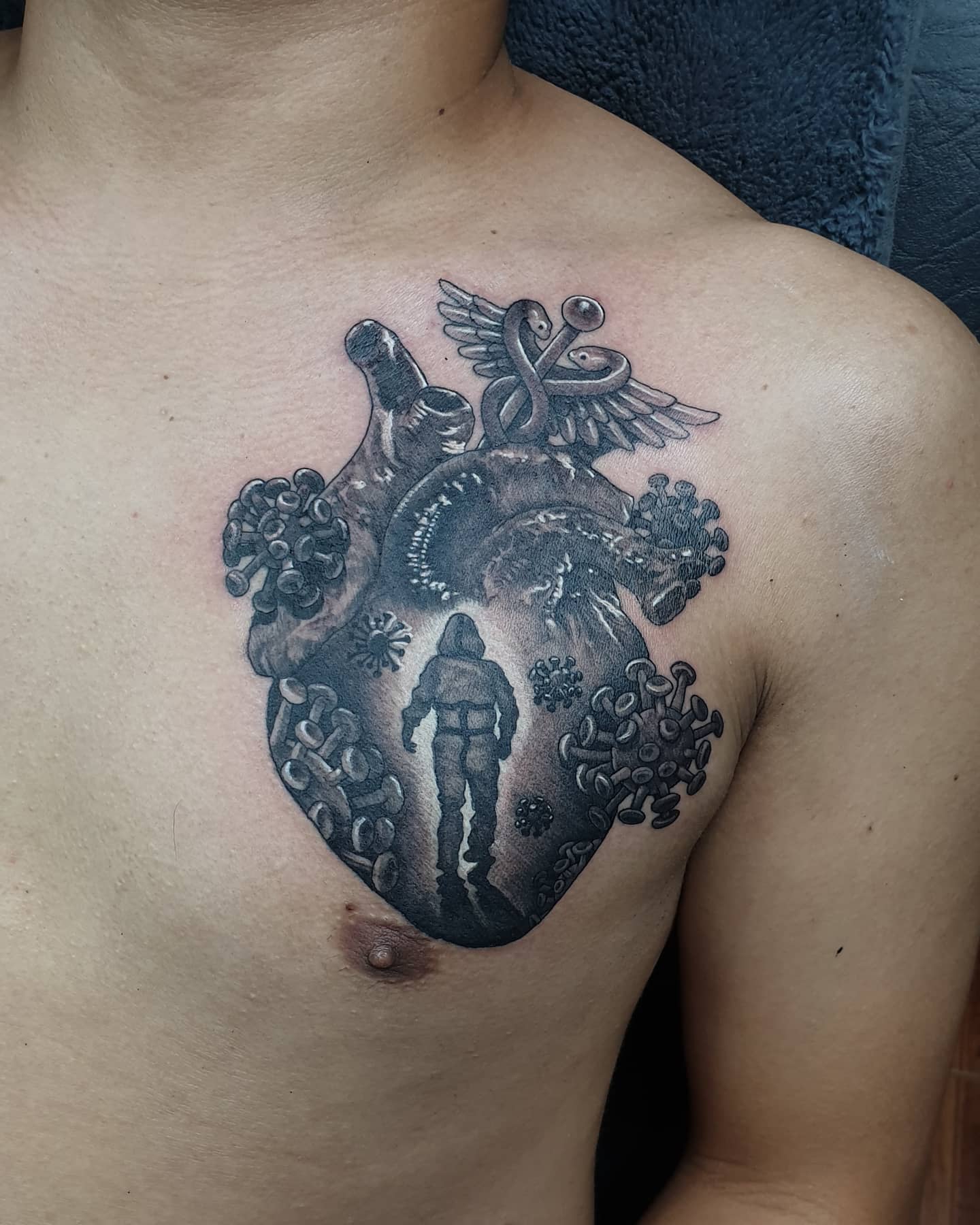 A black and white tattoo of a heart, a winged staff, viruses, and an individual on a chest